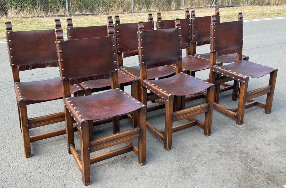A set of eight dark oak dining chairs featuring thick brown leather slung seats and backrests trimmed with brass studs and leather fringe. View III