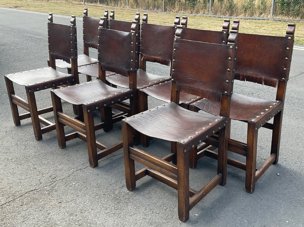 A set of eight dark oak dining chairs featuring thick brown leather slung seats and backrests trimmed with brass studs and leather fringe. View IV