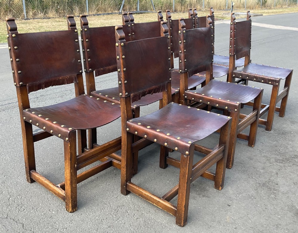 A set of eight dark oak dining chairs featuring thick brown leather slung seats and backrests trimmed with brass studs and leather fringe. View V