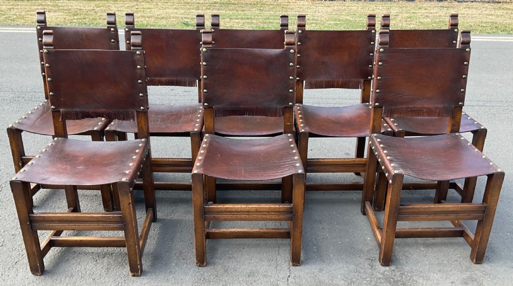 A set of eight dark oak dining chairs featuring thick brown leather slung seats and backrests trimmed with brass studs and leather fringe. View VI