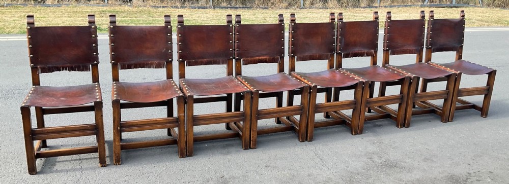 A set of eight dark oak dining chairs featuring thick brown leather slung seats and backrests trimmed with brass studs and leather fringe. View VII