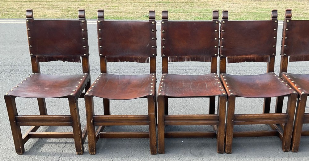 A set of eight dark oak dining chairs featuring thick brown leather slung seats and backrests trimmed with brass studs and leather fringe. View VIII