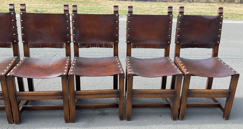 A set of eight dark oak dining chairs featuring thick brown leather slung seats and backrests trimmed with brass studs and leather fringe. View IX