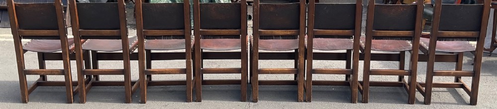 A set of eight dark oak dining chairs featuring thick brown leather slung seats and backrests trimmed with brass studs and leather fringe. View X