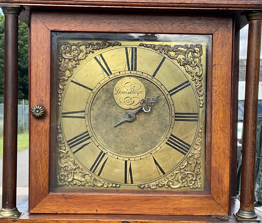 An antique Georgian oak longcase clock featuring a square brass dial, turned hood columns, and a tall wooden trunk door. View V