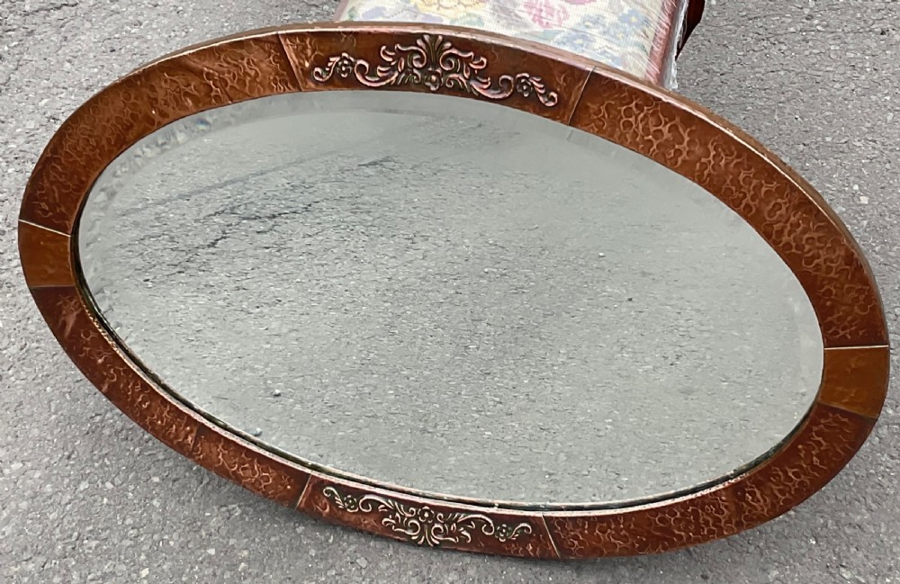 An oval Arts and Crafts wall mirror featuring a planished copper frame decorated with embossed floral relief motifs surrounding a bevelled glass plate.