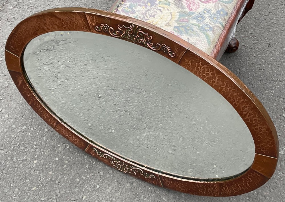 An oval Arts and Crafts wall mirror featuring a planished copper frame decorated with embossed floral relief motifs surrounding a bevelled glass plate. View II