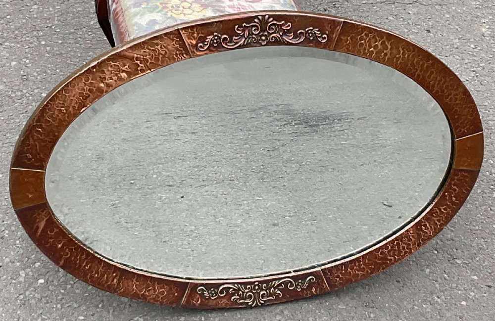 An oval Arts and Crafts wall mirror featuring a planished copper frame decorated with embossed floral relief motifs surrounding a bevelled glass plate. View III