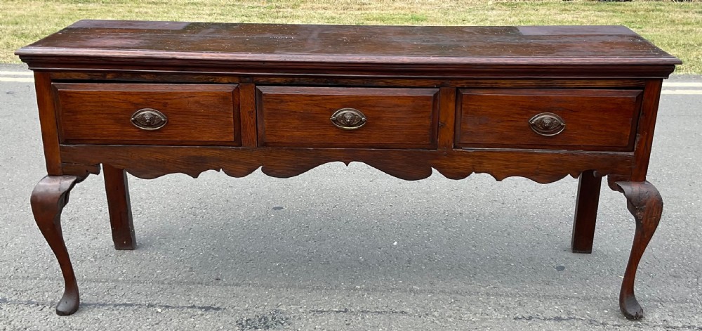 A Georgian dark oak dresser base featuring three drawers with oval brass handles, a shaped apron, and front cabriole legs.