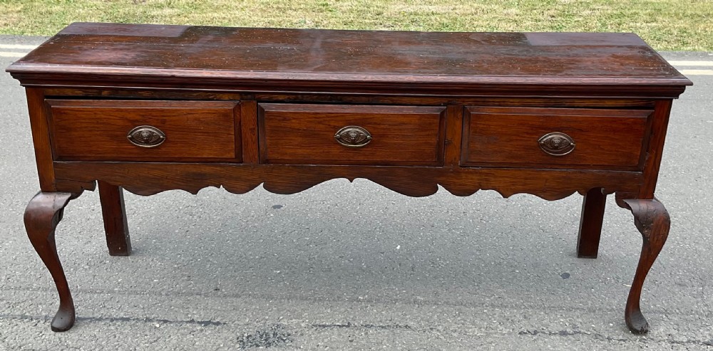 A Georgian dark oak dresser base featuring three drawers with oval brass handles, a shaped apron, and front cabriole legs. View III