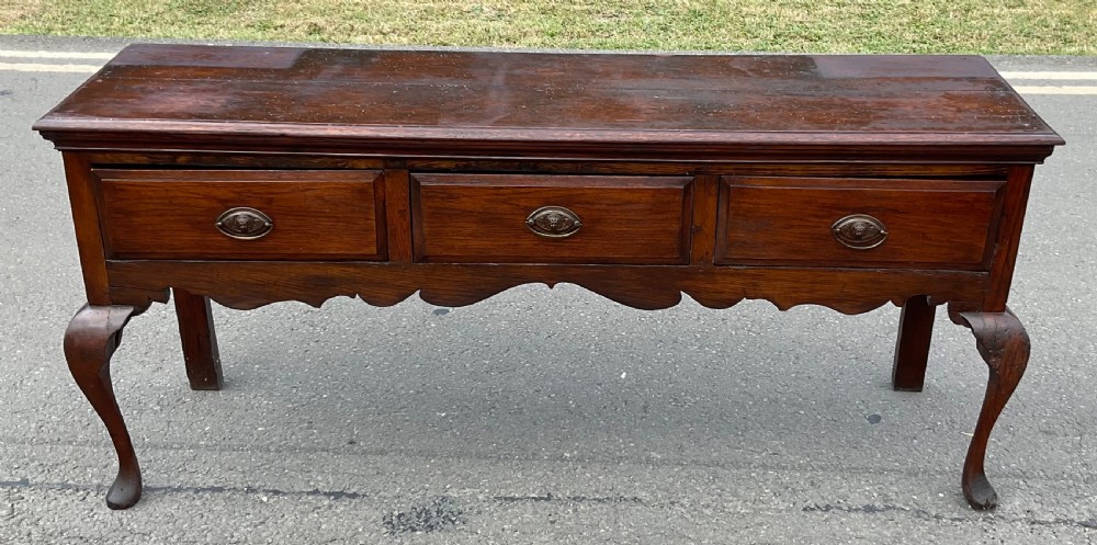 A Georgian dark oak dresser base featuring three drawers with oval brass handles, a shaped apron, and front cabriole legs. View IV
