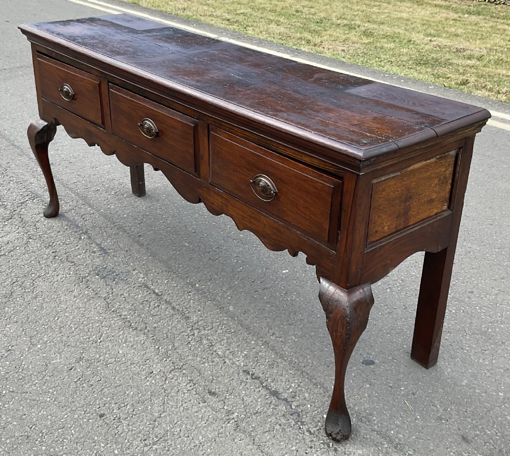 A Georgian dark oak dresser base featuring three drawers with oval brass handles, a shaped apron, and front cabriole legs. View VI