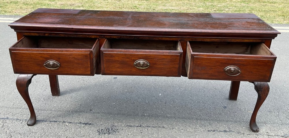 A Georgian dark oak dresser base featuring three drawers with oval brass handles, a shaped apron, and front cabriole legs. View X