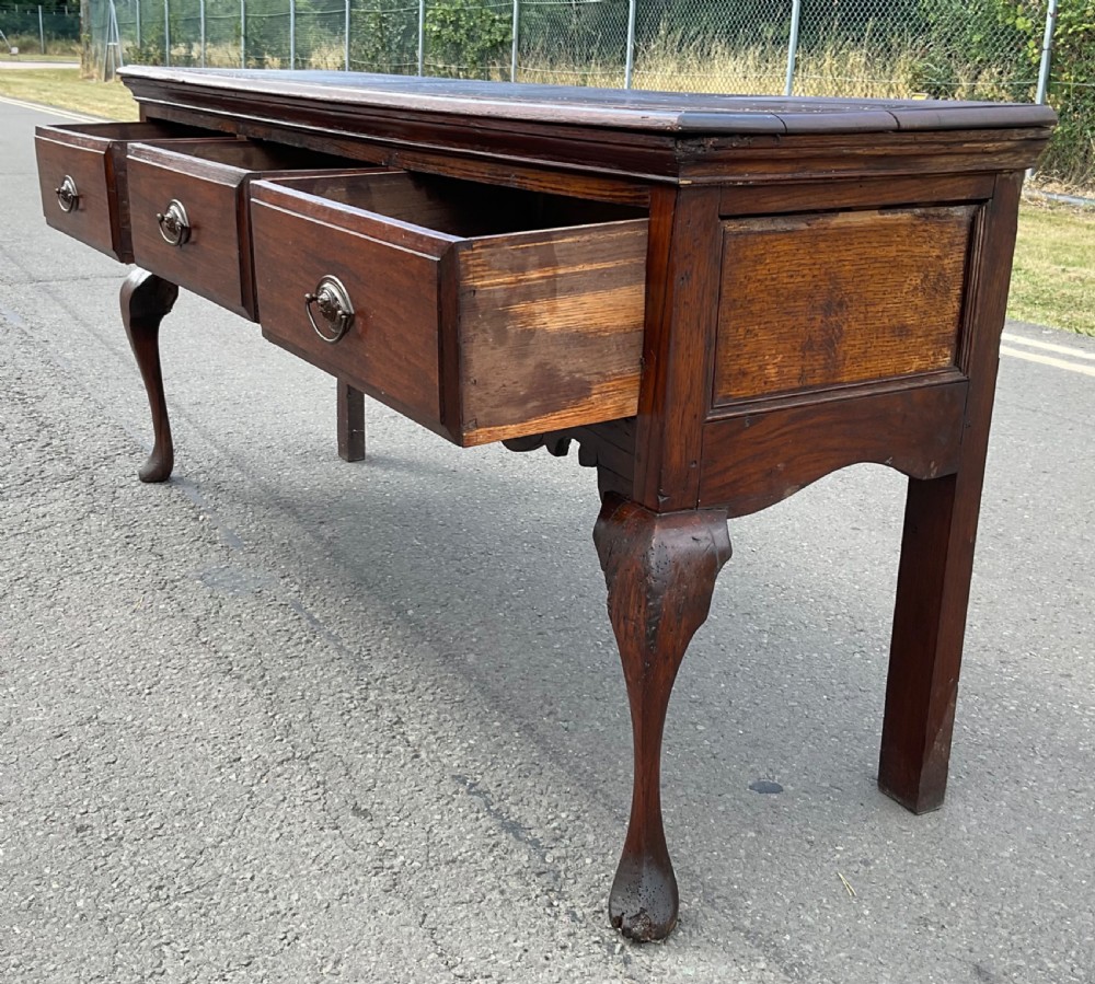 A Georgian dark oak dresser base featuring three drawers with oval brass handles, a shaped apron, and front cabriole legs. View XI