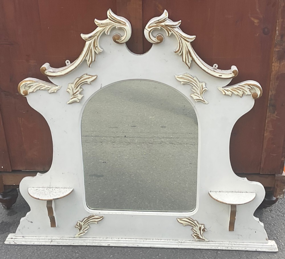 An Edwardian cream painted overmantel mirror with an arched glass centre, carved gold accent flourishes, and two small side display shelves.
