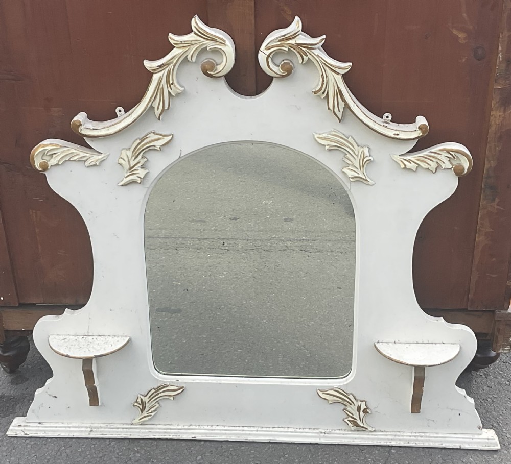 An Edwardian cream painted overmantel mirror with an arched glass centre, carved gold accent flourishes, and two small side display shelves. View II