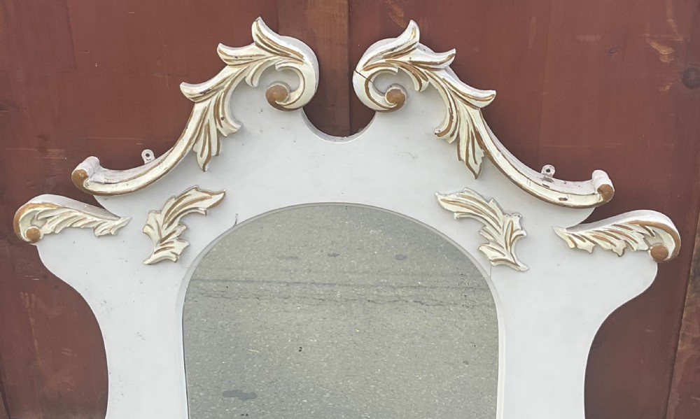 An Edwardian cream painted overmantel mirror with an arched glass centre, carved gold accent flourishes, and two small side display shelves. View IV