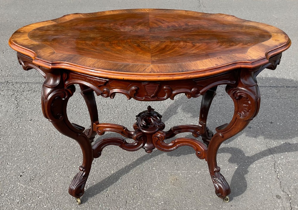 A Victorian mahogany centre table featuring a serpentine oval top with bookmatched veneer, carved cabriole legs, a central X-stretcher with finial, and brass castors.