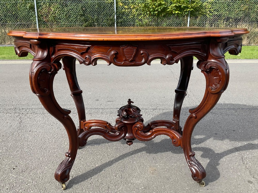 A Victorian mahogany centre table featuring a serpentine oval top with bookmatched veneer, carved cabriole legs, a central X-stretcher with finial, and brass castors. View II
