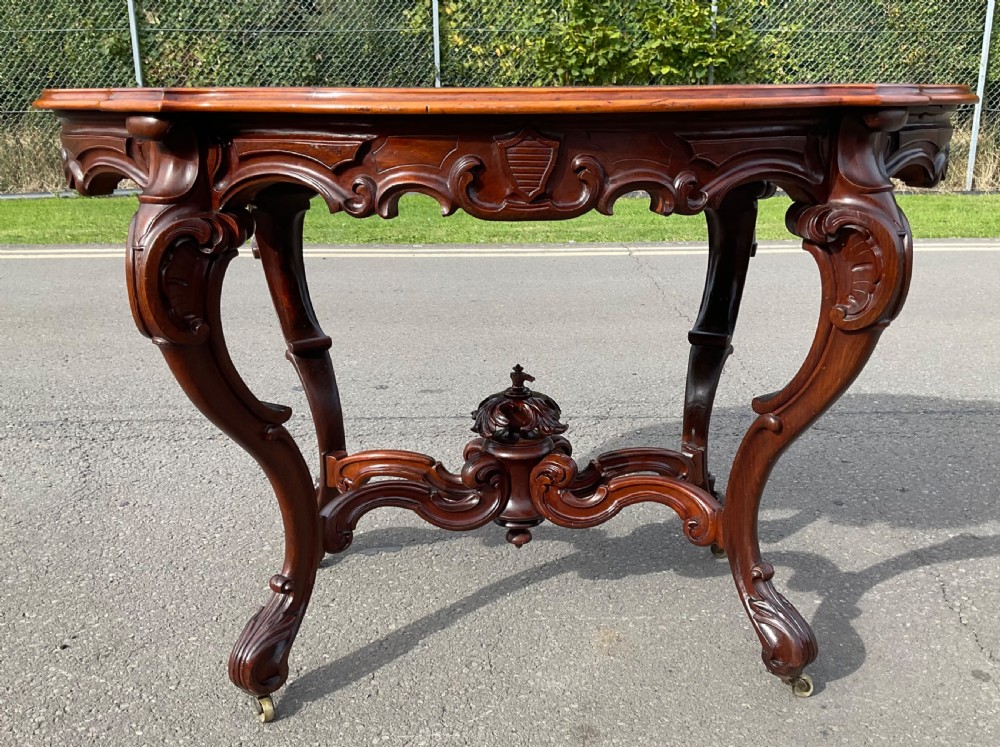 A Victorian mahogany centre table featuring a serpentine oval top with bookmatched veneer, carved cabriole legs, a central X-stretcher with finial, and brass castors. View III