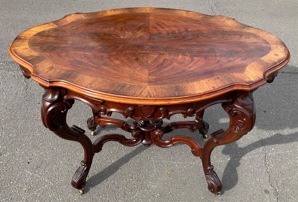 A Victorian mahogany centre table featuring a serpentine oval top with bookmatched veneer, carved cabriole legs, a central X-stretcher with finial, and brass castors. View V