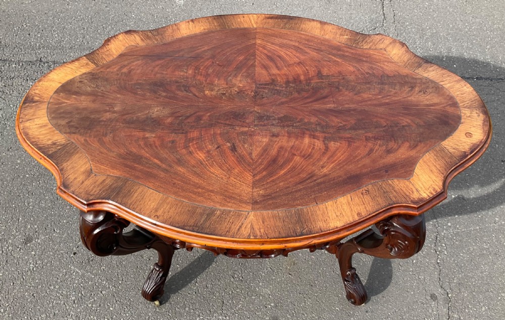 A Victorian mahogany centre table featuring a serpentine oval top with bookmatched veneer, carved cabriole legs, a central X-stretcher with finial, and brass castors. View VI