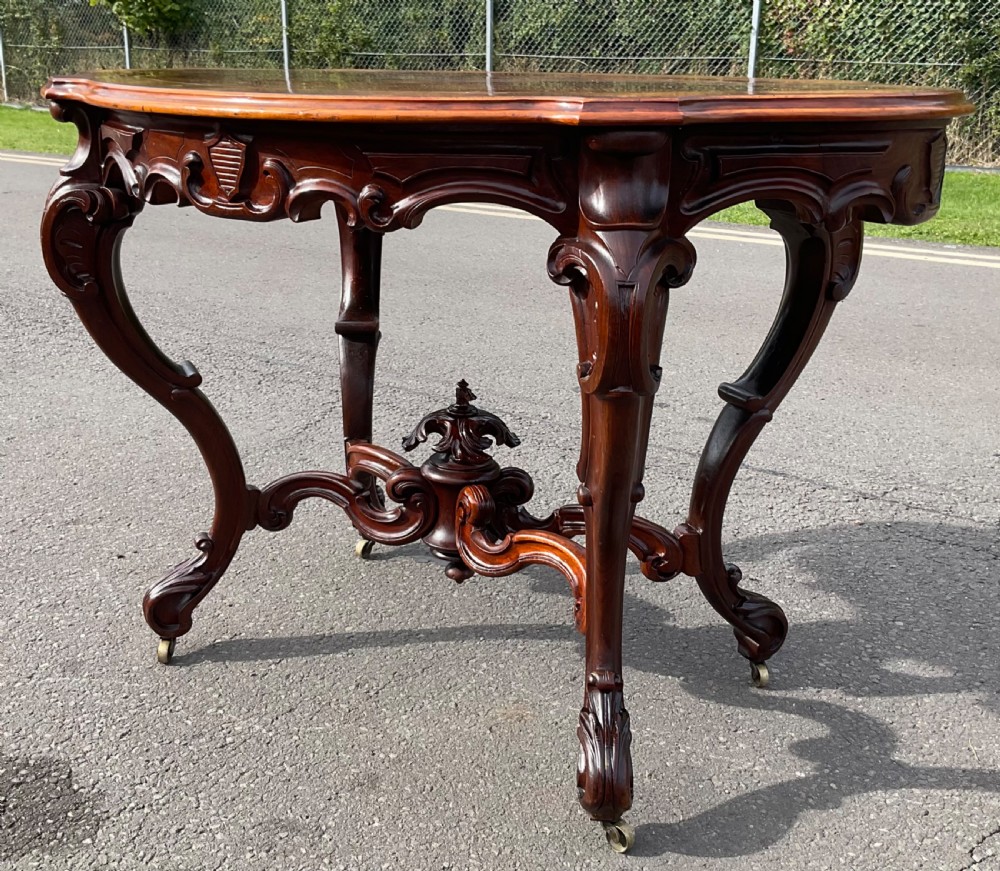 A Victorian mahogany centre table featuring a serpentine oval top with bookmatched veneer, carved cabriole legs, a central X-stretcher with finial, and brass castors. View VII