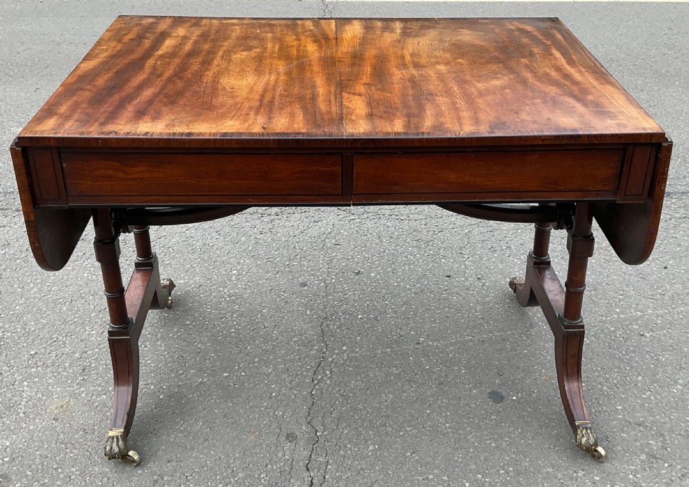 A Georgian flame mahogany drop-leaf sofa table featuring two frieze drawers, twin pedestal sabre legs, and brass paw feet with castors.