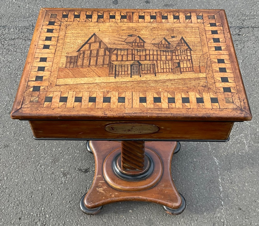 A Victorian wooden pedestal table featuring an inlaid pictorial top depicting a timber-framed building framed by a geometric border, supported by a spiral-turned stem on a platform base.
