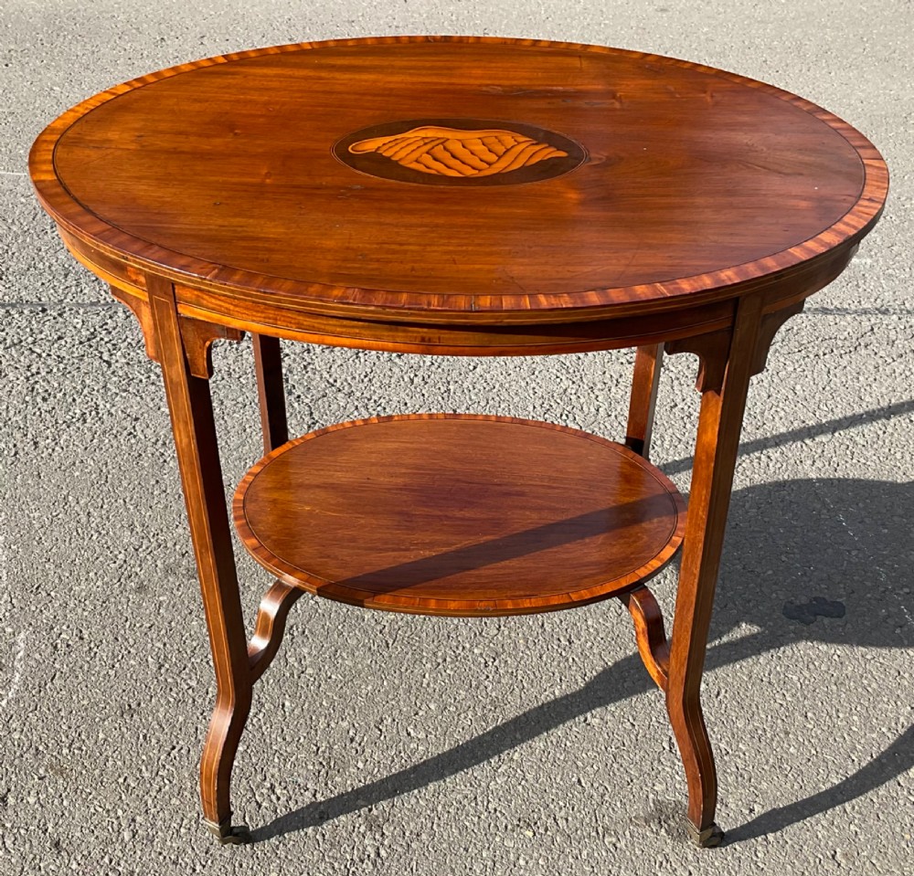 An Edwardian oval mahogany centre table featuring a central shell marquetry inlay, satinwood crossbanded top, lower tier shelf, and splayed legs on brass castors.