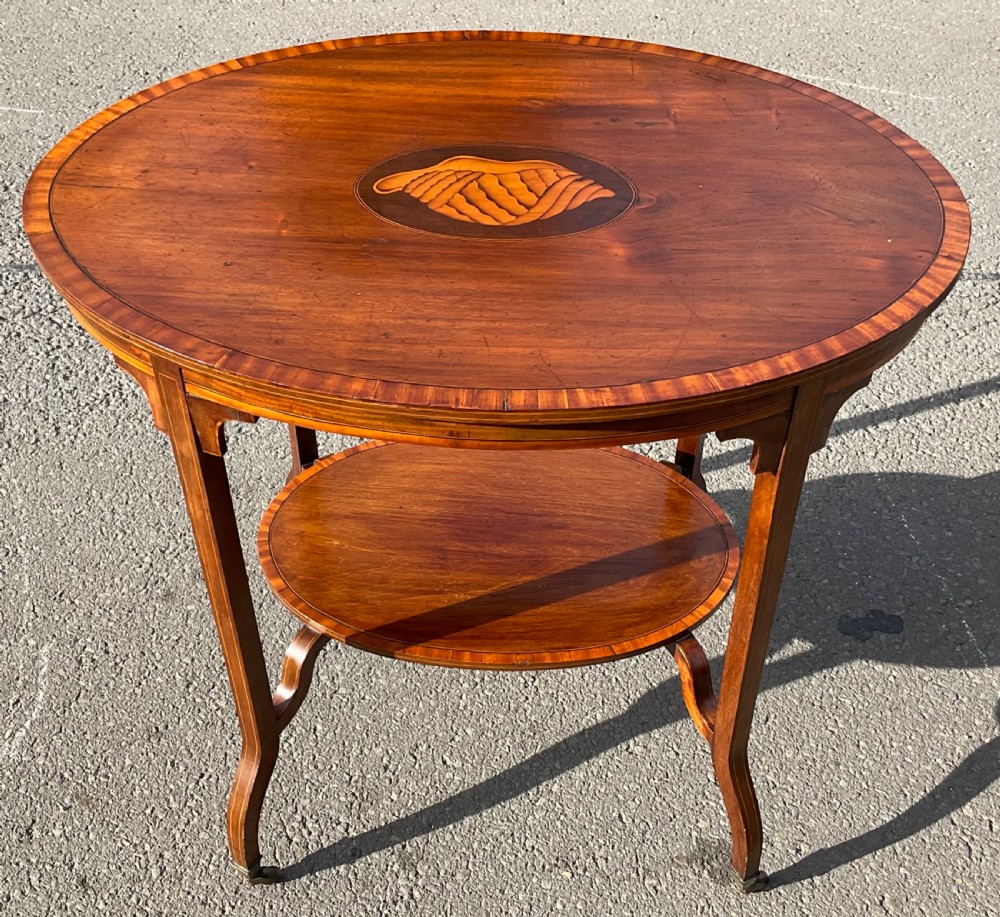 An Edwardian oval mahogany centre table featuring a central shell marquetry inlay, satinwood crossbanded top, lower tier shelf, and splayed legs on brass castors. View III