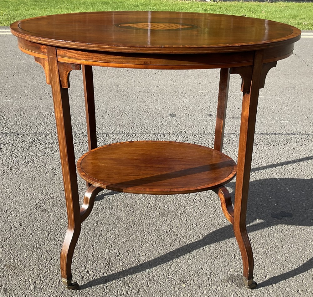 An Edwardian oval mahogany centre table featuring a central shell marquetry inlay, satinwood crossbanded top, lower tier shelf, and splayed legs on brass castors. View VI