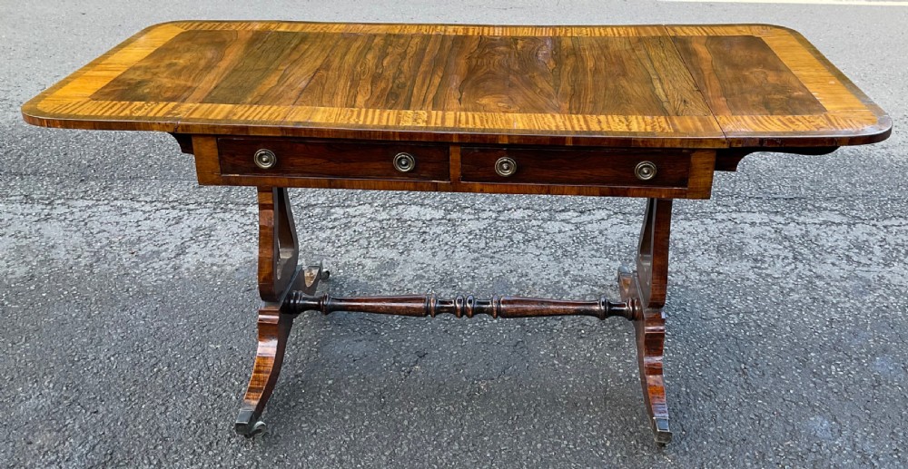 A Georgian walnut drop-leaf sofa table with crossbanded top inlay, two frieze drawers with brass ring handles, turned stretcher support, and splayed legs on castors. View X