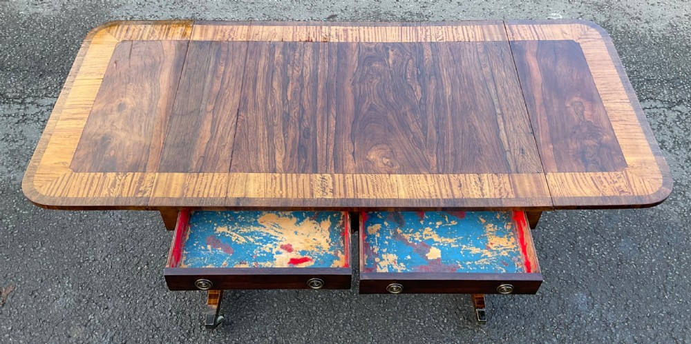 A Georgian walnut drop-leaf sofa table with crossbanded top inlay, two frieze drawers with brass ring handles, turned stretcher support, and splayed legs on castors. View XII