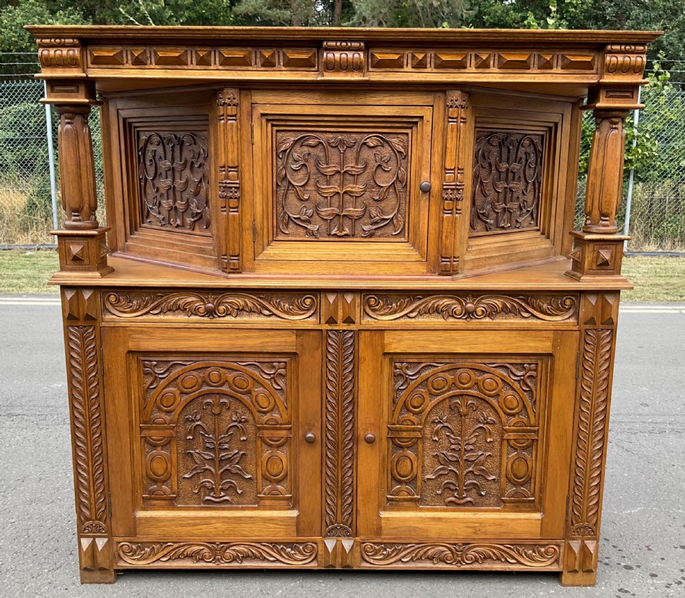 A golden oak court cupboard featuring deeply carved foliate door panels, turned support columns, and geometric moulding.