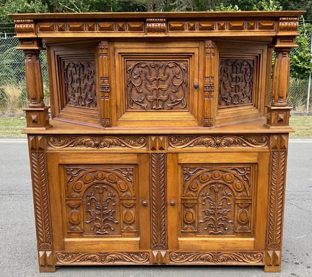 A golden oak court cupboard featuring deeply carved foliate door panels, turned support columns, and geometric moulding. View II