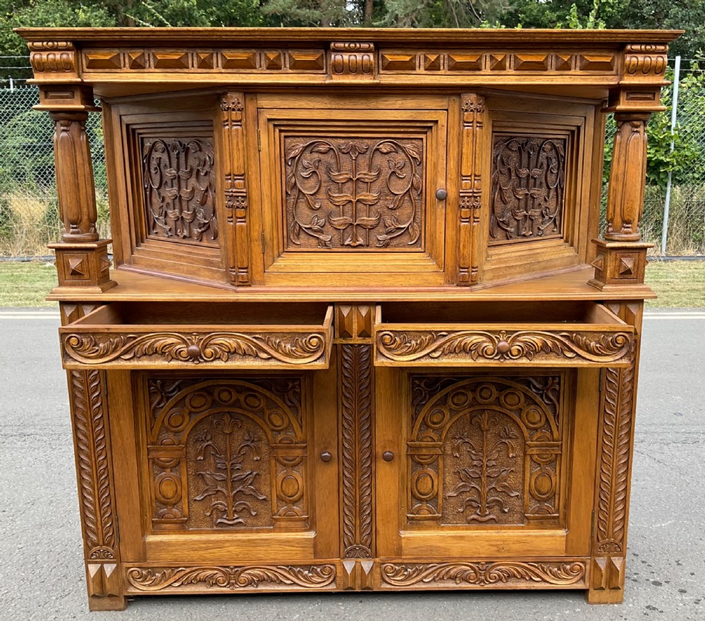 A golden oak court cupboard featuring deeply carved foliate door panels, turned support columns, and geometric moulding. View VII