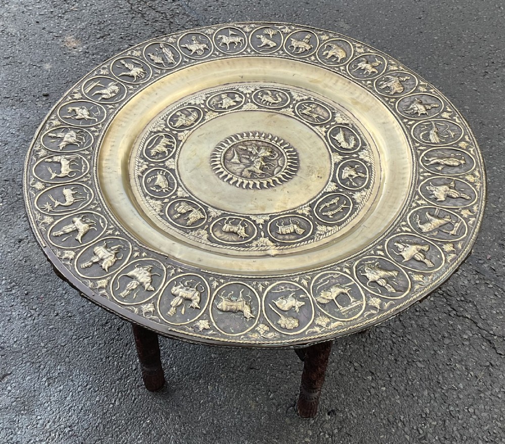 A circular low coffee table with an embossed brass platter top featuring zodiac symbols resting on dark carved wooden legs.