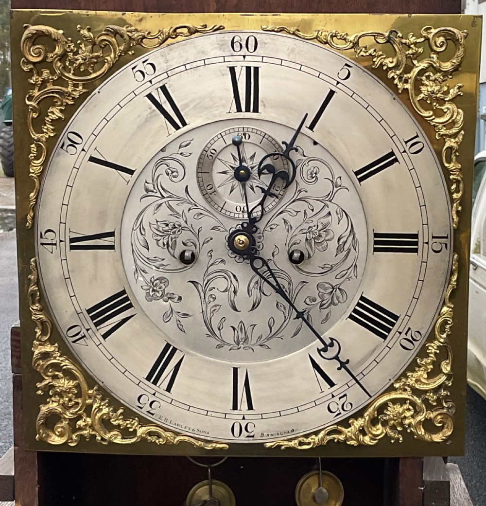 A tall carved dark oak longcase clock featuring a carved trunk door and a brass and silvered dial behind a glass hood window. View VII