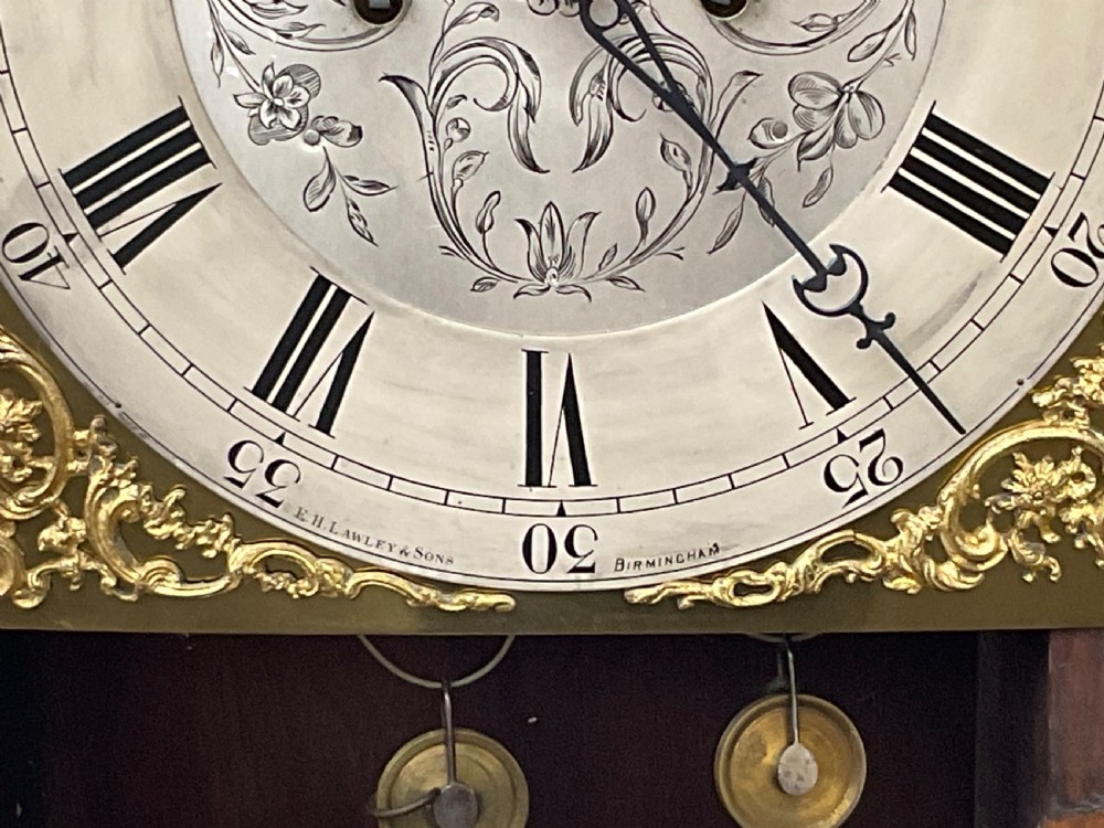 A tall carved dark oak longcase clock featuring a carved trunk door and a brass and silvered dial behind a glass hood window. View IX