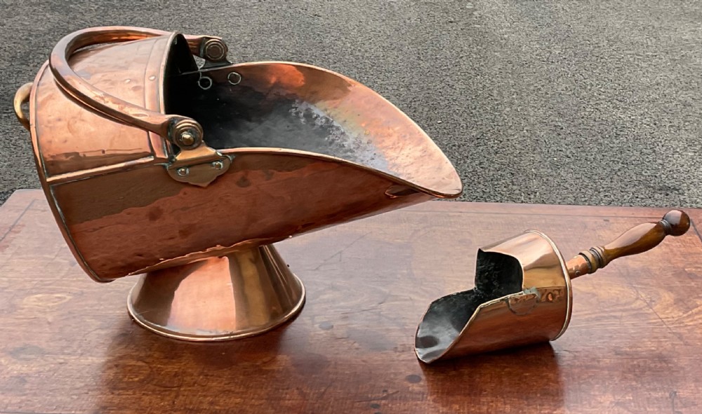A Victorian helmet-shaped copper coal hod on a pedestal base with a swing handle alongside a matching copper scoop with a turned wooden handle.