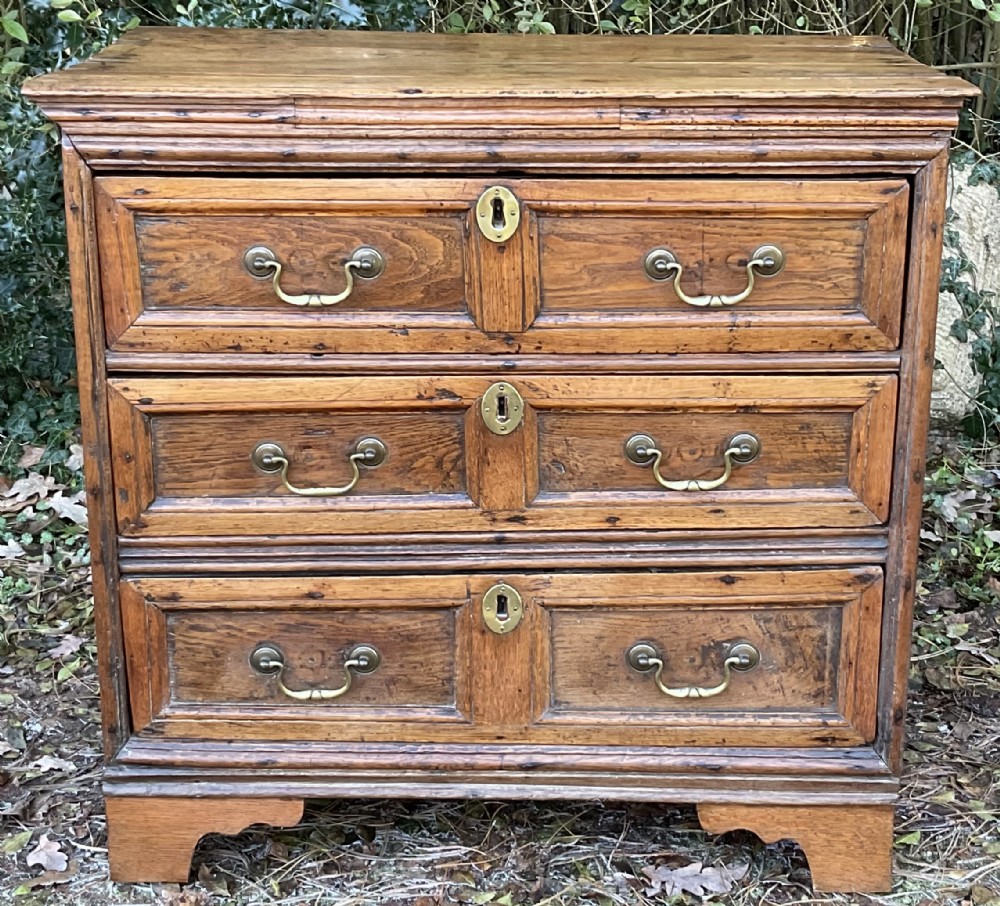 An early oak chest of drawers featuring three geometric panelled drawer fronts with brass drop handles and keyhole escutcheons.