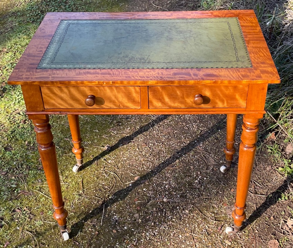 A Victorian writing table made of golden figured wood featuring a green leather insert top, two drawers with turned knobs, and turned legs on ceramic castors.