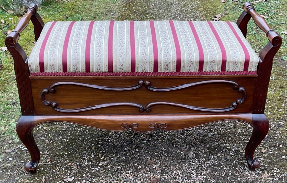 An antique Edwardian mahogany duet piano stool featuring turned side handles, a red and beige striped upholstered seat, carved scrollwork apron, and cabriole legs.
