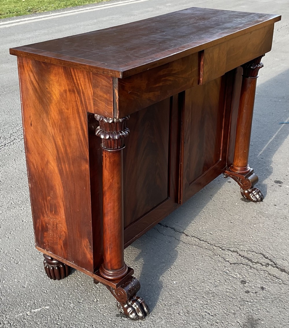 A William IV flame mahogany chiffonier base featuring carved column pillars, lion paw front feet, two frieze drawers, and panelled cupboard doors.