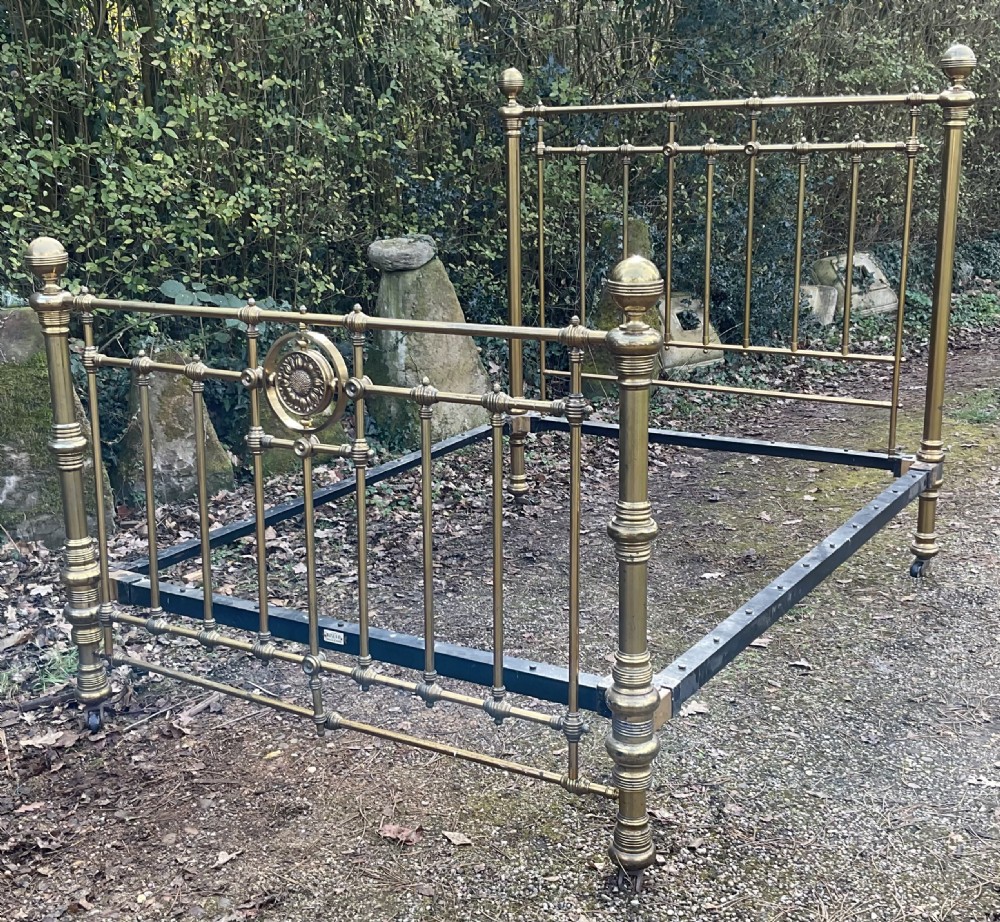 An antique brass five-foot bed frame featuring turned spindles, a central sunflower rosette motif on the footboard, spherical finials, and black iron side rails on castors.