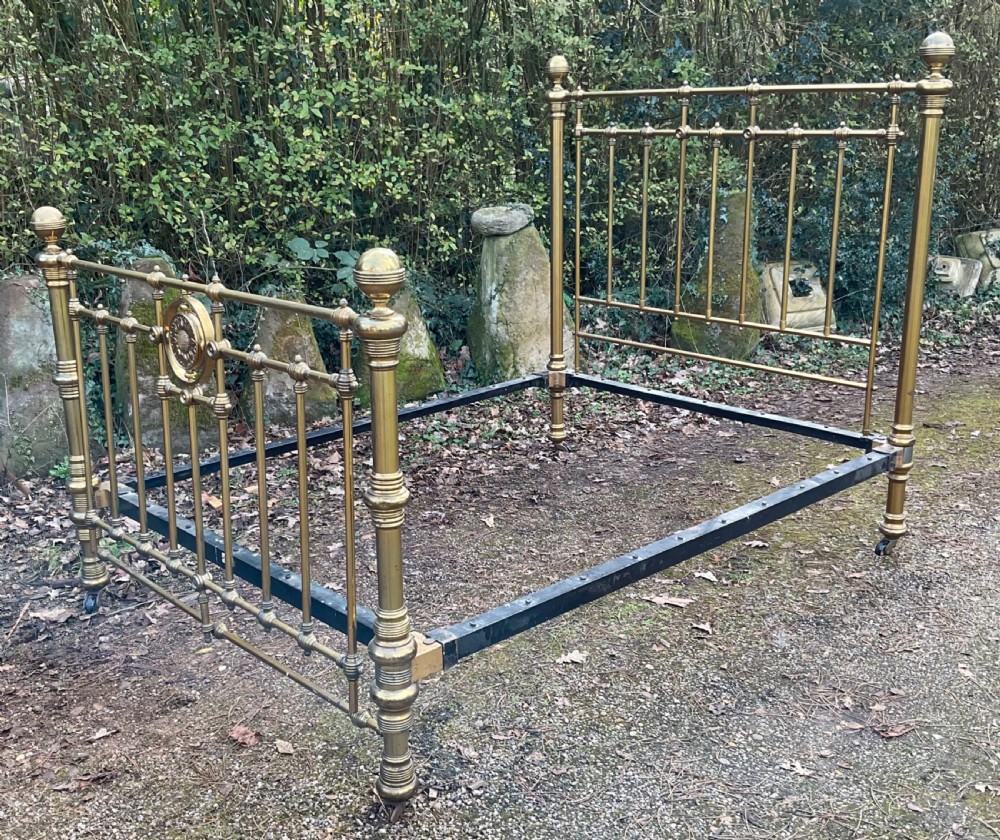 An antique brass five-foot bed frame featuring turned spindles, a central sunflower rosette motif on the footboard, spherical finials, and black iron side rails on castors. View III