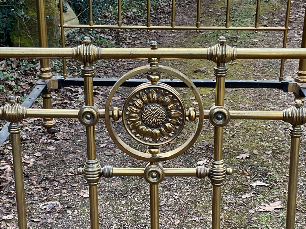An antique brass five-foot bed frame featuring turned spindles, a central sunflower rosette motif on the footboard, spherical finials, and black iron side rails on castors. View V