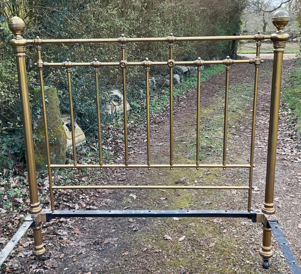 An antique brass five-foot bed frame featuring turned spindles, a central sunflower rosette motif on the footboard, spherical finials, and black iron side rails on castors. View VI