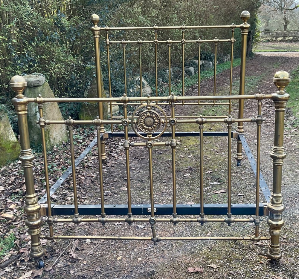 An antique brass five-foot bed frame featuring turned spindles, a central sunflower rosette motif on the footboard, spherical finials, and black iron side rails on castors. View VII